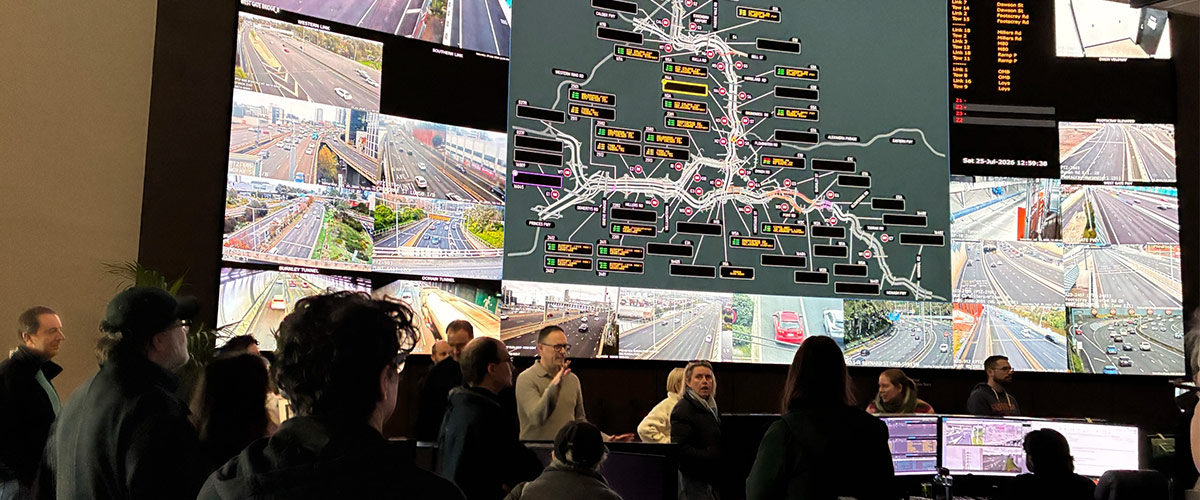 A group of people stand in front of a large screen, there is a road network on the screen.
